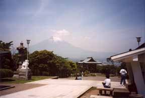 A photo of Fujisan (Mt. Fuji) in the far distance, on a hazy summer
 day, viewed from a relaxed position within an open, paved area of the
 grounds of a modern Peace Park Buddhist temple in Japan.