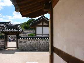 A photo of a few
 traditional structures in an old upperclass housing compound historical park
 area in Seoul, Korea; visible in the distance, through a roofed gateway in
 a low stone wall, is a teacher photographing a small group of young
 children.